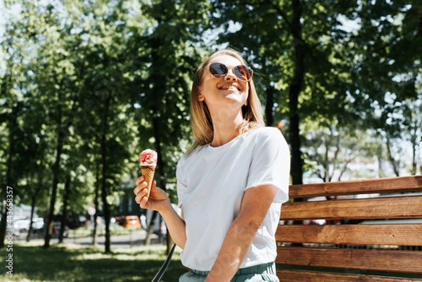 Obraz Joyful young woman relaxing on a park bench looking up enjoying the sun and summer weather. Cute smiling girl eating ice cream in a waffle cone, outdoors. Summer lifestyle