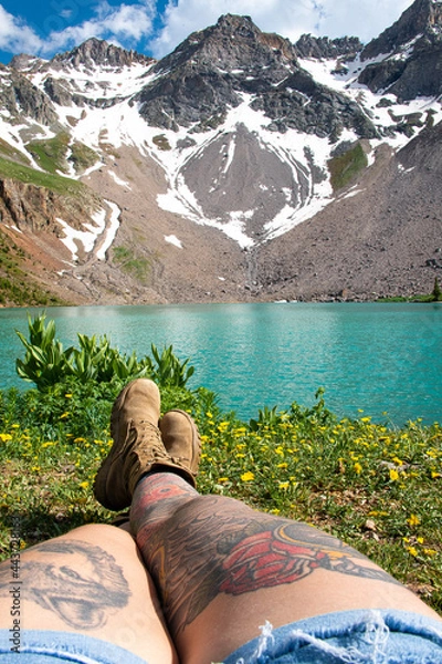 Fototapeta View of a woman's legs with tattoos who is sitting in front of a mountain peak and lake. The view is of Blue lake in Colorado. 