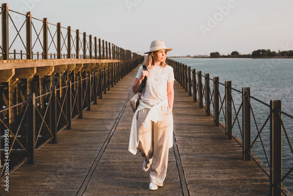 Obraz A girl in light clothes and a hat is walking along the pier and looking at the sunset against the backdrop of the seascape. Huelva, Spain.