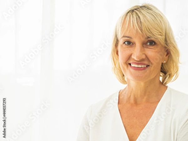 Fototapeta Portrait of beautiful middle-aged woman smiling friendly and looking in camera in the living room. Realistic images without retouching with their imperfections..