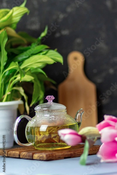 Fototapeta green tea with rose petals in a transparent teapot on a wooden table on a black background. The background is blurred. Tea drinking.