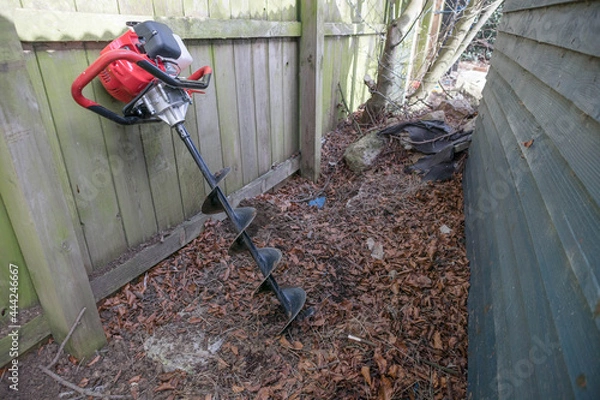 Fototapeta Drill like excavation hole digger power tool leaning against a fence waiting to be used to dig fence post holes