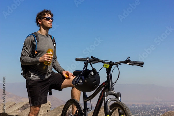 Fototapeta Young biker with sunglasses holding water bottle next to mountain bike on sunny day. Handsome man taking a break while and enjoying splendid views of Santiago city in Chile