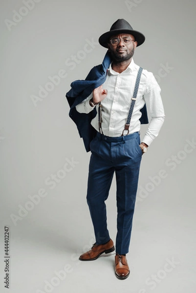 Fototapeta Full-length photo. Attractive stylish african american businessman in a hat and with suspenders holds a jacket with his hand on his shoulder
