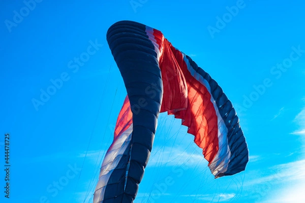Fototapeta Red-white-blue paraglider filled with the wind close-up. A multi-colored wing of a paraglider against a clear sky. Paraglider in the sunlight against the blue sky.