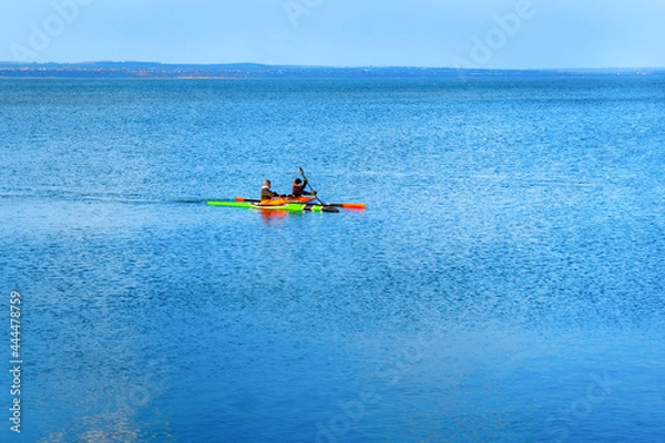 Fototapeta A company of young people on kayaks is training at sea. A pair of multi-colored canoes sail in the blue sea. Spring canoeing guy and girl in the lake