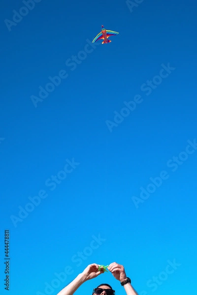 Fototapeta man holds a rainbow color kite flying high in the blue sky in summer. young guy launches a kite in summer in hot weather