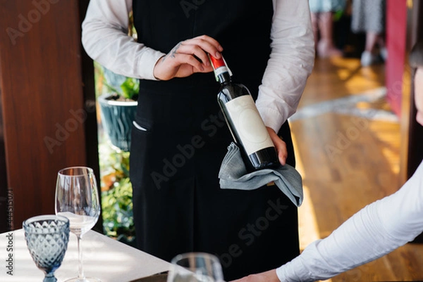Fototapeta A young waiter in a stylish apron demonstrates and offers a fine wine to a client in a restaurant. Customer service.