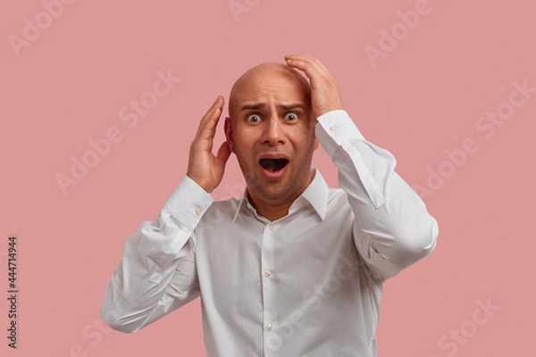 Fototapeta Scared bald man with bristle being in panic, keeps hands near face, trying to cover himself, sees something terrible, has horror opened mouth. Dressed in white shirt. Isolated over pink background.