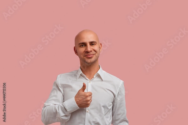 Fototapeta Good work. Portrait of happy bald man with bristle keeps thumb raised, being in good mood, shows his confirmation. Satisfied with something. Dressed in white shirt. Isolated over pink background.