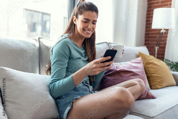 Obraz Pretty young smiling woman using mobile phone while drinking cup of coffee sitting on a couch at home.