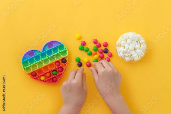 Fototapeta Use the popular pop it toy as a chocolate mold. Children is hands put colorful candies in a popit heart-shaped silicone mold on a yellow background. Step-by-step photo instructions. Step 2.