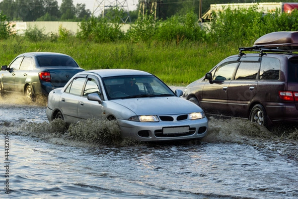 Fototapeta The car is driving through a deep puddle. Splashes are flying from under the wheels of the car. Flood in the city. The consequences of a downpour.