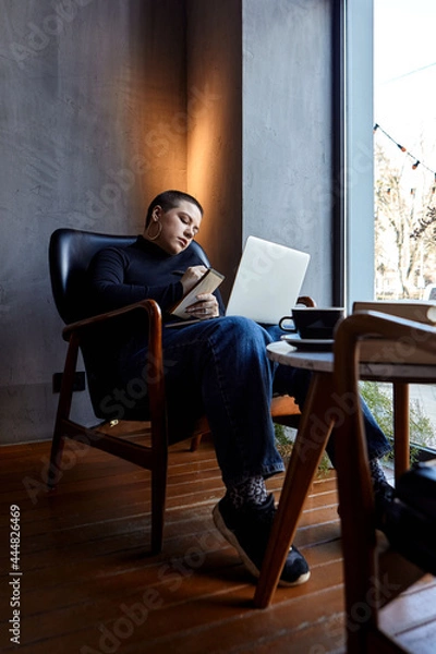 Fototapeta A young girl with glasses, with short hair is sitting in a cafe and working on a laptop. The girl looks at her laptop and makes notes in a notebook. The concept of distance learning