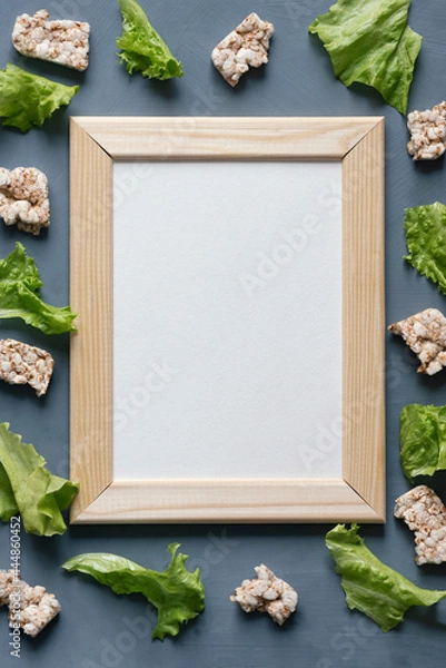 Obraz Stationery mock-up scene dedicated to healthy eating. Blank white greeting card in wooden frame surrounded by whole grain crisps and lettuce pieces on textured gray background.