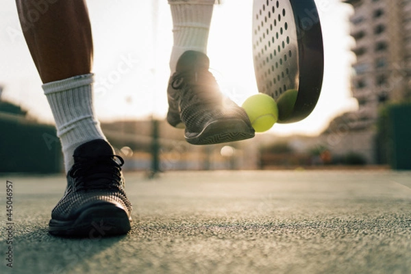 Fototapeta Man lifting paddle ball with his racket and shoe on the green court during training in the sunset