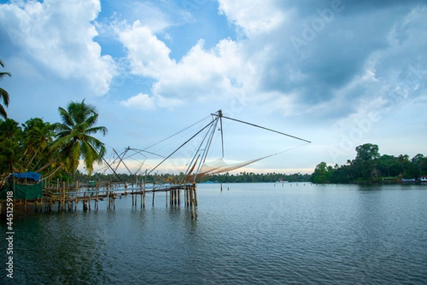 Fototapeta A Chinese fishing net and a small hut with coconut trees under a blue and white cloudy sky, River photography with a Chinese fishing net from Kerala, India