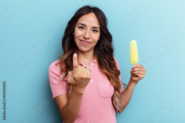 Obraz Young latin woman holding ice cream isolated on blue background pointing with finger at you as if inviting come closer.