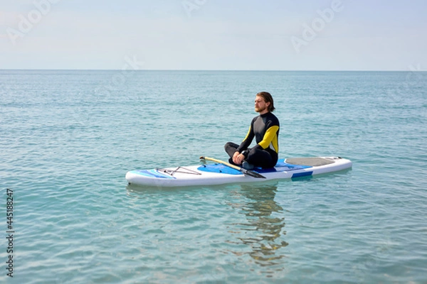 Fototapeta Side view portrait of relaxed bearded guy sitting on paddleboard meditating after sup surfing, in black wetsuit, keep calm, yoga and sport concept. copy space