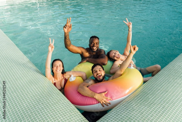 Fototapeta Four multiracial friends with a float in the pool, having fun in summer.