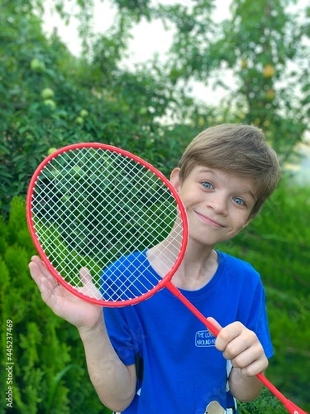 Fototapeta A funny boy with a badminton racket in his hands stands against the background of green fruit trees Children playing in the camp. The child rejoices in summer.