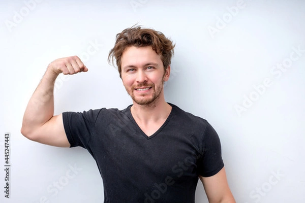 Fototapeta muscular man in casual wear shows muscles on arms, feels proud to be strong and have strength. I am hero. Successful smiling male model raises hands with joy, shows great power. Healthy lifestyle