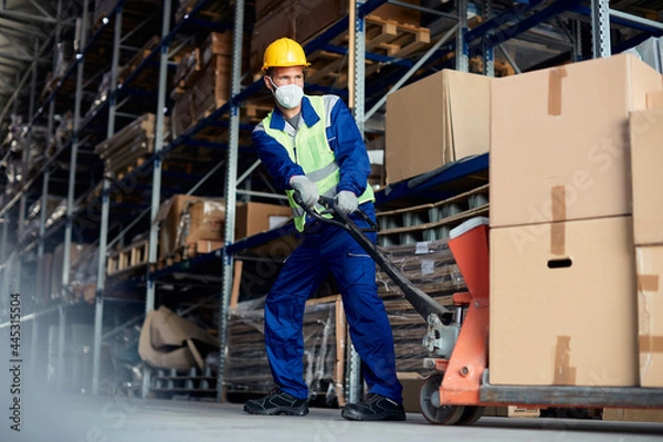 Fototapeta Male worker unloading pallet shipment boxes in warehouse and wearing face mask to due to COVID-19 pandemic.