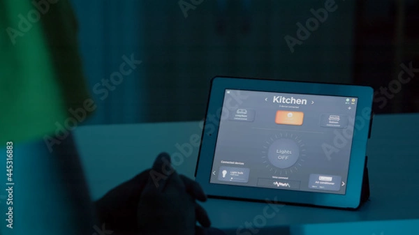Fototapeta Man sitting at desk in smart home automation system talking to voice assistant on tablet to turn on lights of house. Male using intelligent software, gadget placed on kitchen table controlling light