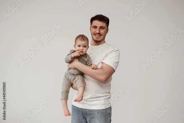 Fototapeta Young educator smiling, happy that he was able to calm the baby, looks into the camera holding the child in his arms, the child's teeth are cut, she holds her finger in her mouth and no longer crying