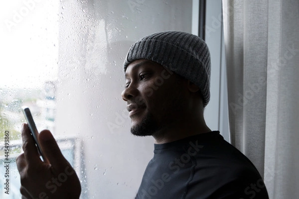 Obraz African American man smiling and looking at his cellphone while standing by the window on a rainy winter day.