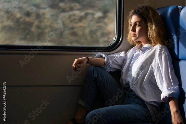 Fototapeta Girl passenger traveling by train sits on the seat and looks out the window