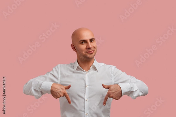 Fototapeta This place os perfect. Horizontal photo of joyful bald man with mysterious smile, points down and shows excellent place to dinner. Dressed in white shirt. Has bristle. Isolated over pink background.