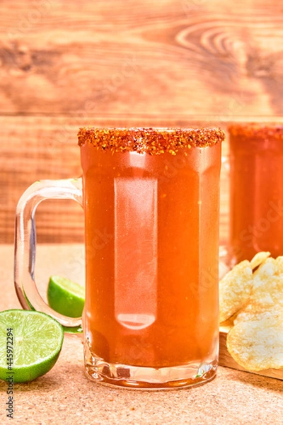Obraz mexican style beer mug (Chelada/Michelada) with lime, hot sauce and chamoy, frosted with chili powder. accompanied by potato chips on a cork surface and wooden background. close up view