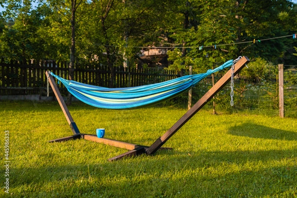 Fototapeta Blue, wooden hammock and blue cup standing on green grass in a rural scenery in the evening sun