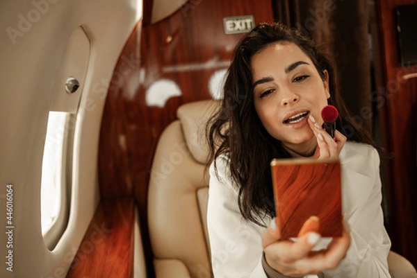 Fototapeta Young brunette in a white suit corrects makeup with a mirror in powder flying aboard a business jet plane