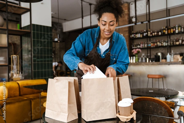 Obraz Black waitress in apron packing orders while working at cafe