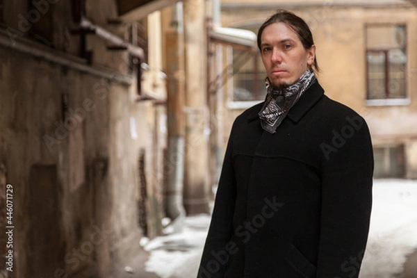 Fototapeta Long-haired guy stands in a snow-covered arch in a black coat