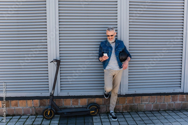 Fototapeta full length of happy and mature man in sunglasses using smartphone near e-scooter