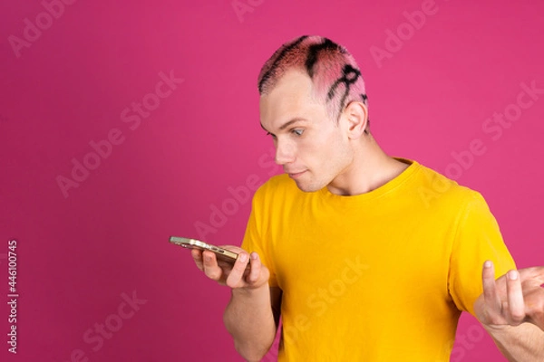 Fototapeta European handsome young man in yellow t shirt on pink background with mobile phone look at screen shrugging shoulder
