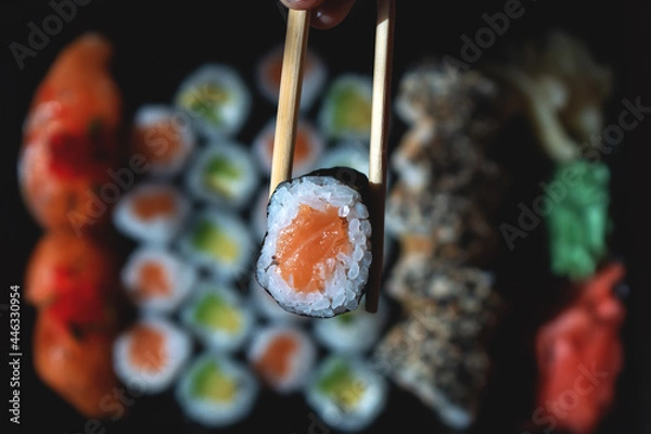 Obraz A girl holds a small roll with chopsticks on the background of a set with rolls
