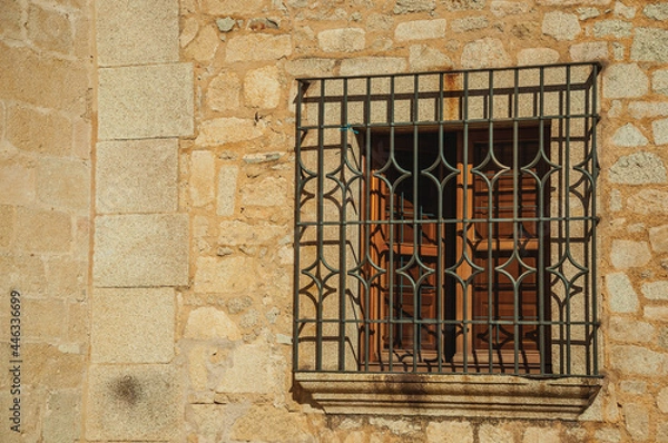 Fototapeta Wooden window with wrought iron grid on the stone facade of gothic building, in a sunny day at Caceres. A fully preserved medieval town in Spain.