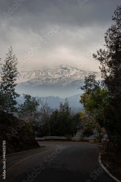 Fototapeta A big mountain with snow in front of a road with trees in a cloudy day