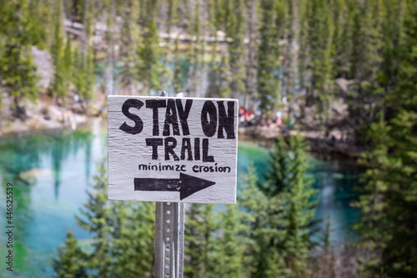 Fototapeta Wooden Sign with a right pointing arrow and Stay on Trail Minimize Erosion text on the Grassi Lakes Hike with blurred trees and turquoise lake in the background