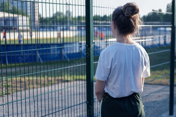 Fototapeta A girl stands and looks through the fence at the athletes playing at the stadium. The girl wants, but cannot play sports.