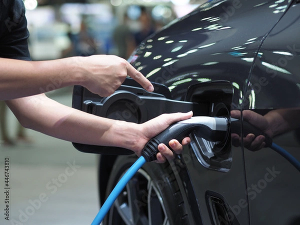 Fototapeta Hand hold and point finger at  electric charging vehicle plug and light flare for recharge battery of car black color clean energy power for future concept