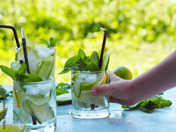 Fototapeta A child is hand takes a summer cold drink with ice, cucumber, lemon and lime to cool in the heat.