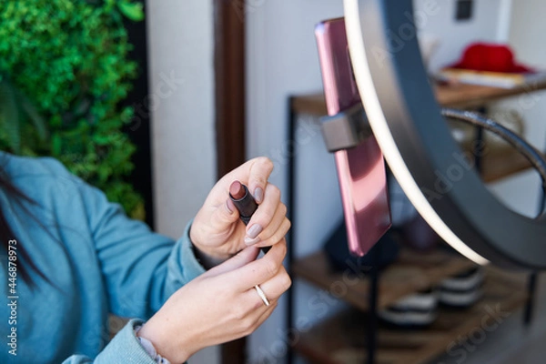 Obraz unrecognizable woman showing her lipstick in front of a tripod with circular light and her cell phone. female influencer blogger live in front of her smartphone. social media concept