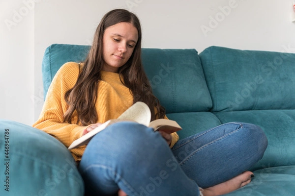 Fototapeta girl reading a book sitting on the sofa