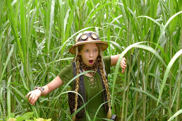 Fototapeta A young girl is dressed up as an explorer. She 
is seen in a tropical jungle environment Her 
face shows an expression of astonishment as 
she observes the jungle area.