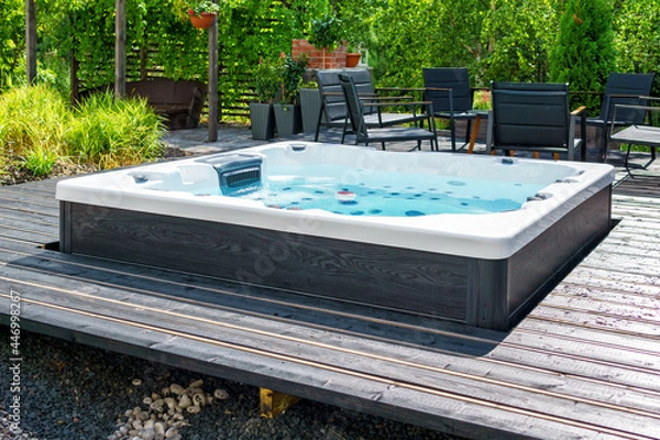 Fototapeta Large hot tub embedded in the backyard terrace. A sunny summer's day in the shelter of a green garden. Everyday luxury and relaxation in your own backyard. Spa complex, vacation and traveling concept.